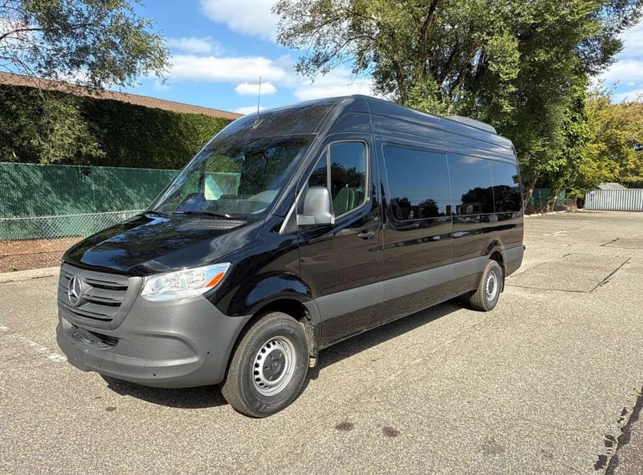 Black high-roof Mercedes-Benz Sprinter passenger van in an empty parking lot, trees behind, front three-quarter view