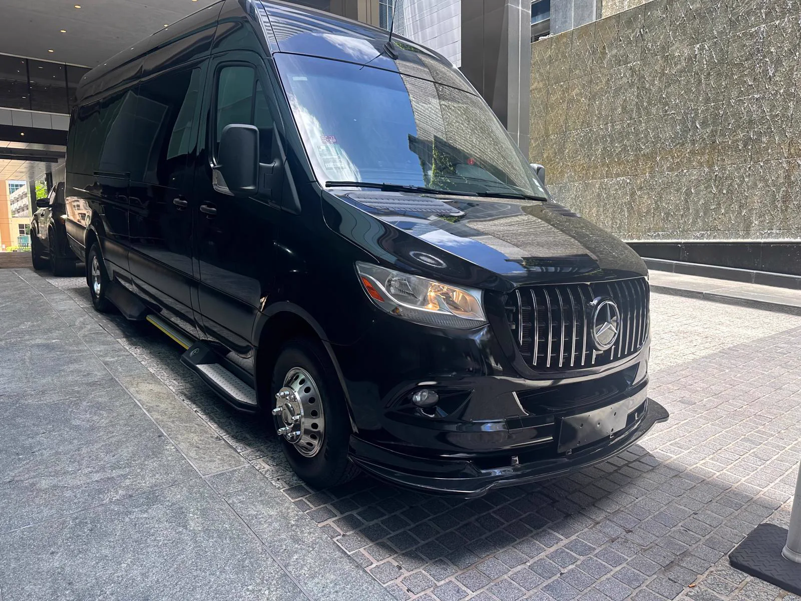 Black Mercedes-Benz Sprinter with chrome wheels parked in daylight at the entrance of a modern building