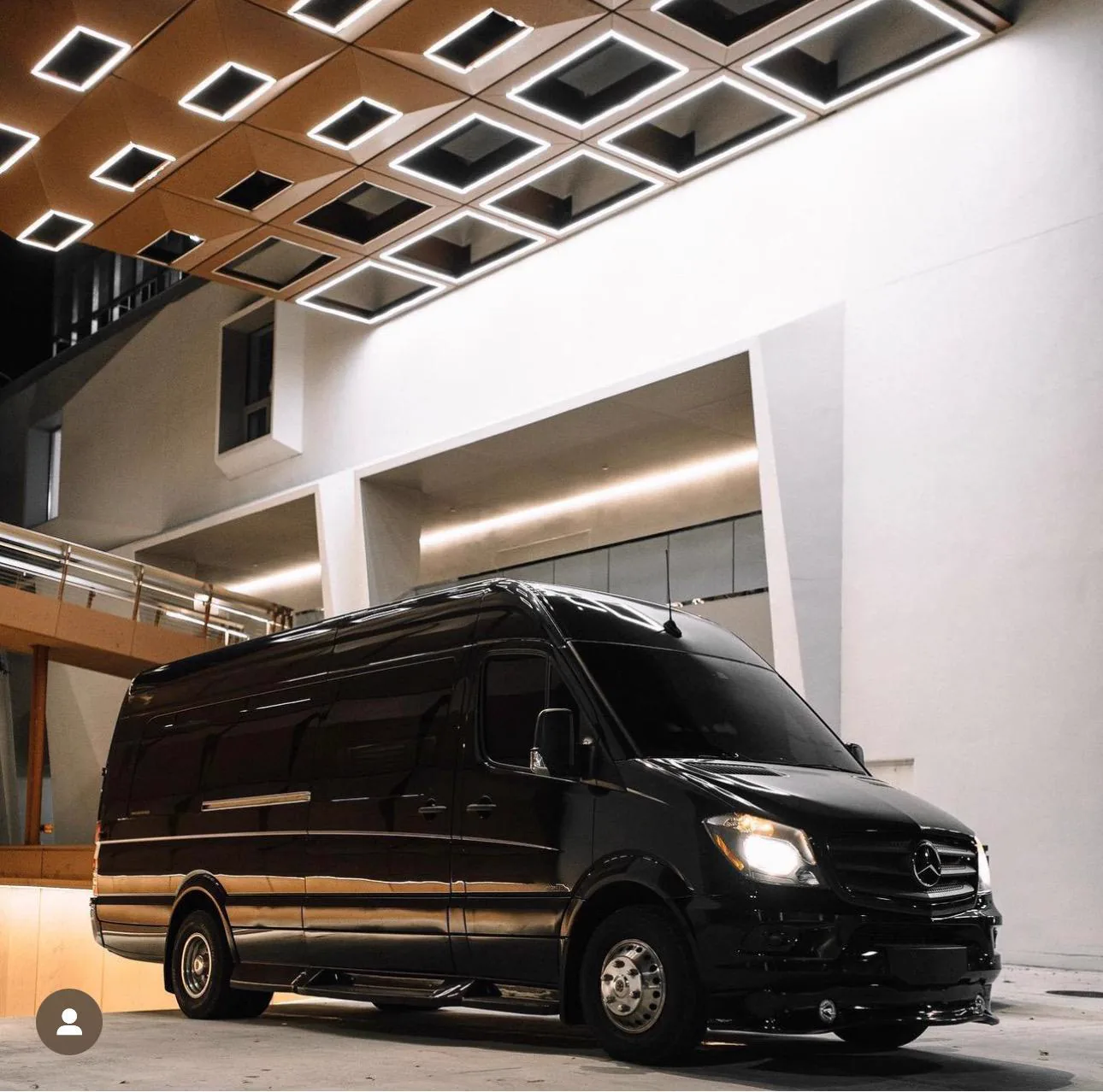 Black Mercedes-Benz Sprinter parked at night at a modern building entrance under a lit coffered ceiling