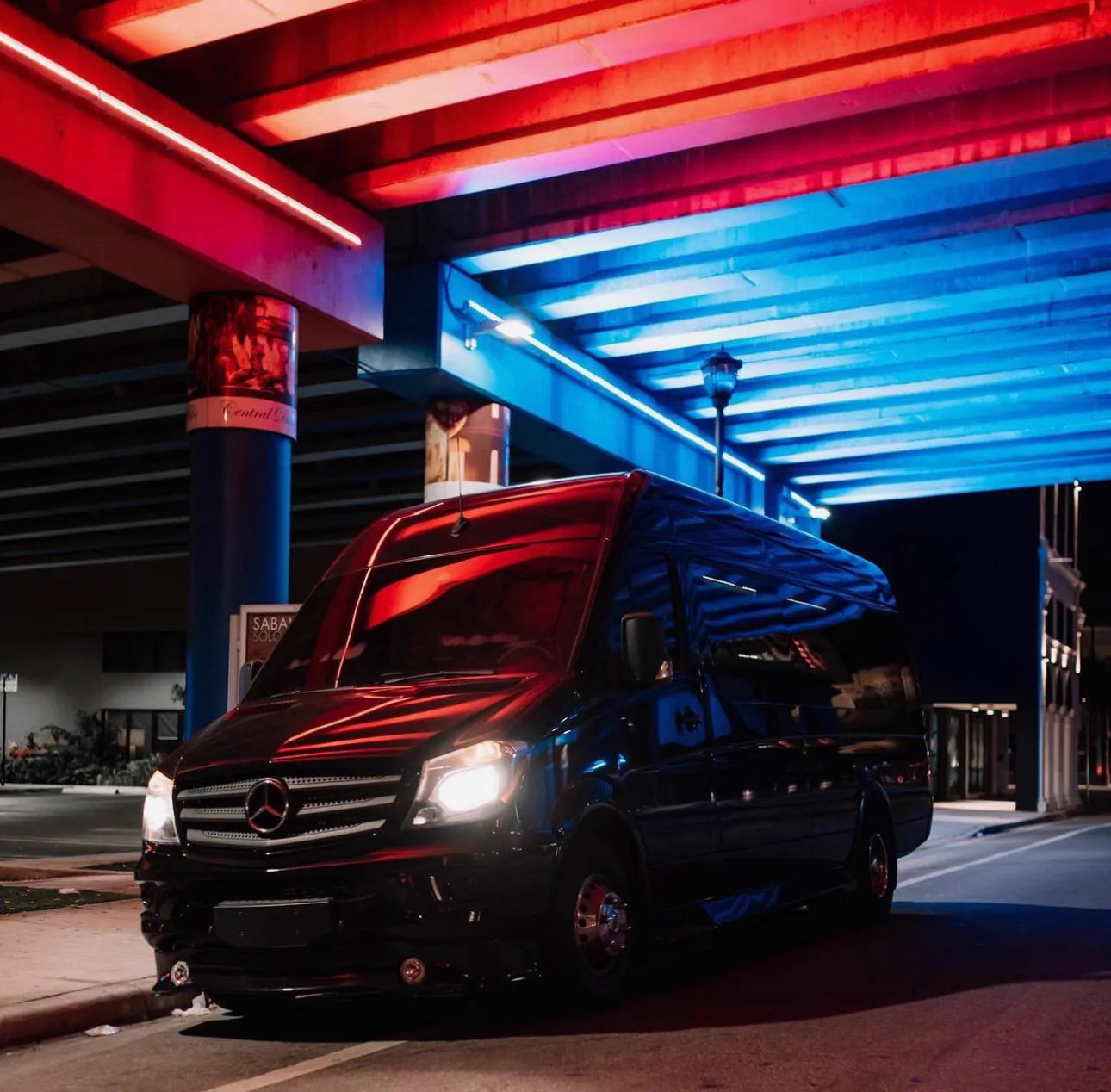 Black Mercedes-Benz Sprinter at night with headlights on beneath red and blue neon strip lighting